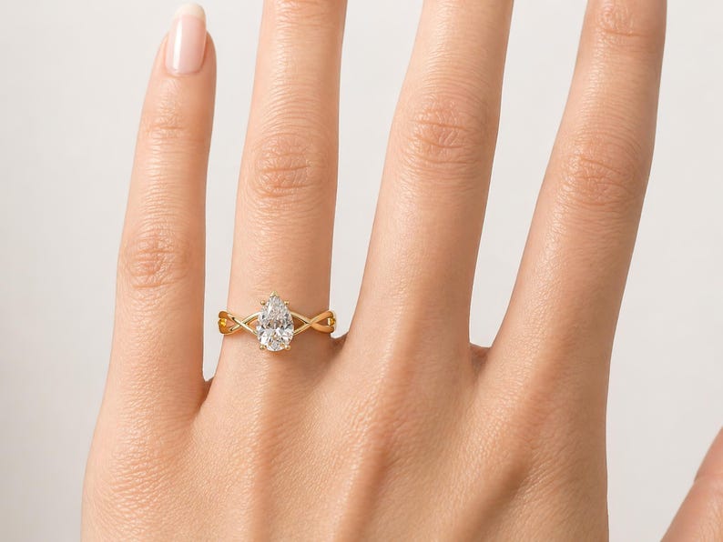 May include: A gold ring with a pear-cut diamond centre stone. The band has an interwoven design. The ring is shown on a person's hand, highlighting the ring's intricate details and sparkle.