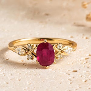 May include: A gold ring featuring a central oval-cut deep red ruby gemstone. The band is accented with four small round-cut diamonds and two marquise-cut diamonds arranged in a symmetrical pattern on either side of the ruby. The brand name Sapheena is visible in the bottom right corner.