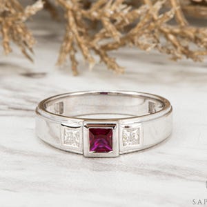 May include: A silver ring featuring a central square, deep red gemstone, with two smaller, square, clear gemstones either side. The ring has a flat band and a contemporary design, suitable for a special event.