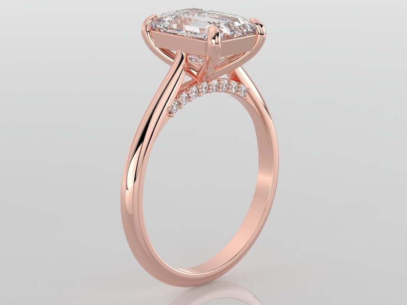 May include: A rose gold engagement ring with a large, clear, square-shaped diamond set in a prong setting. The band is thin and has a row of smaller diamonds set in a pave setting.