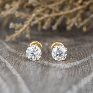 May include: A pair of gold stud earrings with round, clear gemstones. The earrings are set against a textured, brown background.