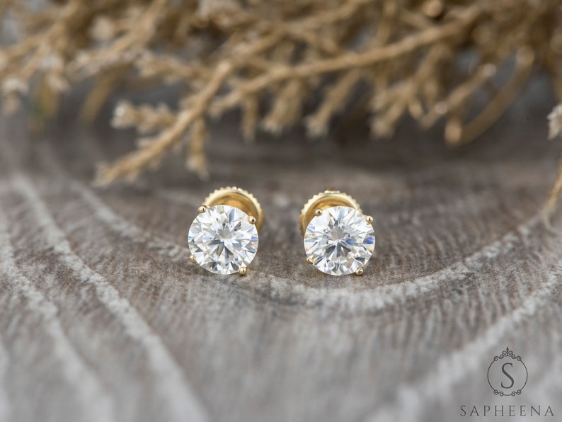 May include: A pair of gold stud earrings with round, clear gemstones. The earrings are set against a textured, brown background.