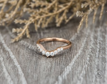 Op de afbeelding: Gebogen ring van roségoud met een rij ronde, heldere edelstenen. De ring is geplaatst tegen een neutrale achtergrond van houtnerf met gedroogd blad in het bovenste deel van de afbeelding. Een delicaat sieraad.