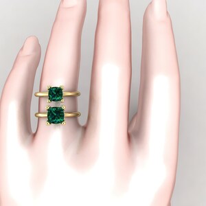Op de afbeelding: Een gouden ring met twee vierkante smaragdgroene edelstenen. De ring zit aan de vinger van een model.