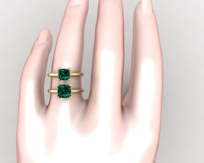 Op de afbeelding: Een gouden ring met twee vierkante smaragdgroene edelstenen. De ring zit aan de vinger van een model.