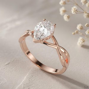 May include: Rose gold engagement ring with a pear-cut diamond. The ring has a twisted band design and a prong setting. The diamond is clear and sparkling, set against a soft, neutral background. A symbol of love and commitment.