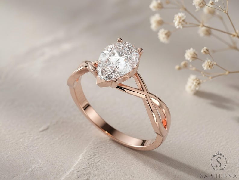 May include: Rose gold engagement ring with a pear-cut diamond. The ring has a twisted band design and a prong setting. The diamond is clear and sparkling, set against a soft, neutral background. A symbol of love and commitment.