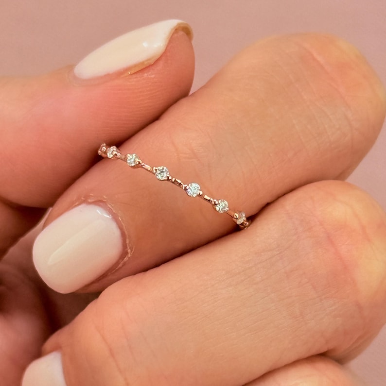 May include: Delicate rose gold band featuring a minimalist design with small, sparkling diamonds arranged in a linear pattern. The diamonds are evenly spaced along the band, creating a subtle yet elegant look. This dainty ring is perfect for everyday wear or special occasions. The metal appears smooth and polished, enhancing the brilliance of the diamonds.