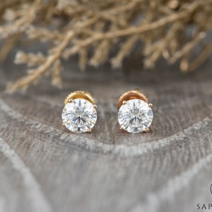 May include: A pair of gold stud earrings with round, clear gemstones. The earrings are set against a backdrop of brown, textured fabric.