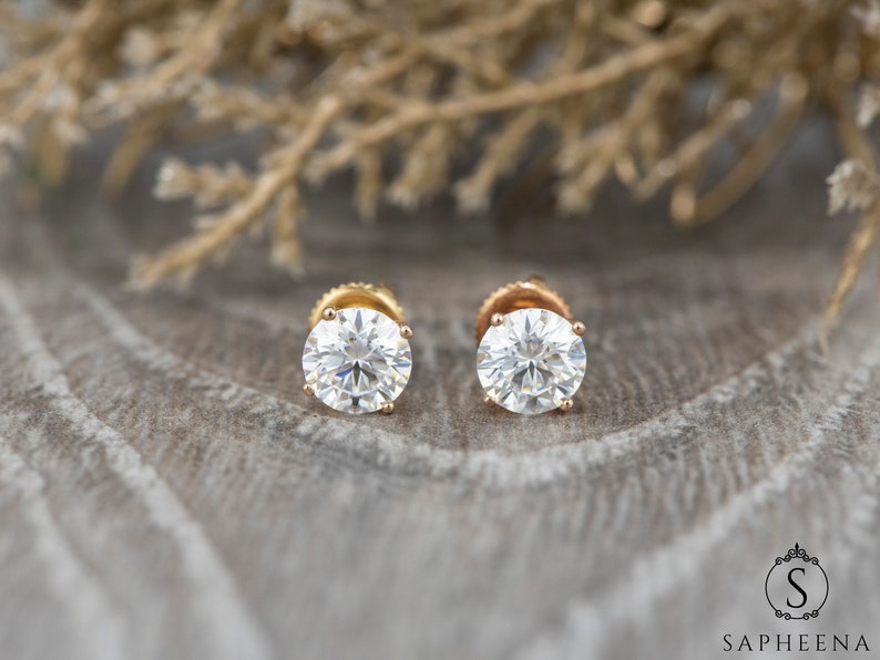 May include: A pair of gold stud earrings with round, clear gemstones. The earrings are set against a backdrop of brown, textured fabric.