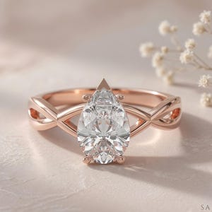 May include: Rose gold engagement ring with a pear-cut diamond. The ring has an infinity-style band with an interwoven design. The diamond is the focal point, reflecting light. The ring is set against a soft, neutral background.