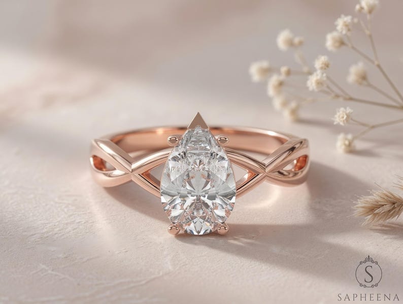 May include: Rose gold engagement ring with a pear-cut diamond. The ring has an infinity-style band with an interwoven design. The diamond is the focal point, reflecting light. The ring is set against a soft, neutral background.