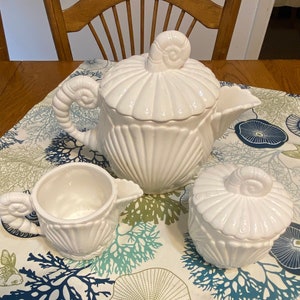 May include: White ceramic teapot, creamer, and sugar bowl set with a seashell design. The teapot has a lid with a seashell finial. The creamer and sugar bowl also have seashell finials.