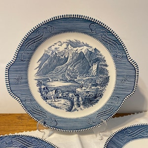 Puede incluir: Un juego de tres platos de cerámica azul y blanco con un diseño vintage. Los platos presentan una escena de paisaje azul y blanco con montañas, árboles y un río. Los platos tienen un borde festoneado y un borde azul y blanco.