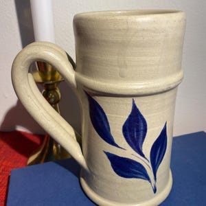 May include: Cream-colored ceramic mug with a large handle and a blue leaf design. The mug has a cylindrical shape with a slightly flared top and a band around the middle. The blue leaves are painted on the side of the mug.