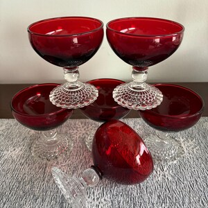 May include: A collection of six ruby red stemmed glasses with a clear, textured base. One glass is lying on its side. The glasses are arranged on a patterned surface, showcasing their vibrant color and elegant design.