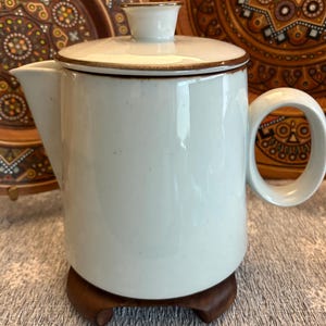 May include: A white ceramic teapot with a lid and handle, featuring a brown trim. The teapot sits on a small, dark wooden stand. Decorative plates with brown and orange patterns are in the background.