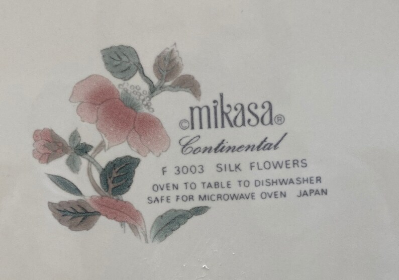 Peut inclure: Vaisselle blanche Mikasa Continental avec un motif floral. Le motif pr&eacute;sente des fleurs roses et rouges avec des feuilles vertes. Le texte "F 3003 SILK FLOWERS" est visible, ainsi que "OVEN TO TABLE TO DISHWASHER SAFE FOR MICROWAVE OVEN JAPAN."