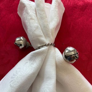 May include: A white fabric napkin folded and secured with a silver-toned metal napkin ring. The ring features a twisted band design and is accented with two polished, spherical jingle bells attached to each side of the central circular band.