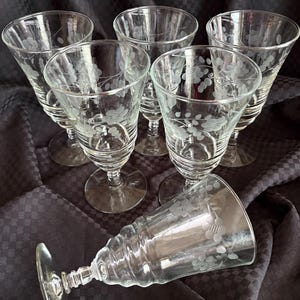 Könnte beinhalten: Sechs klare Weingläser mit geätzten Blumendesigns. Die Gläser haben eine klassische Form mit einer ausgestellten Schale, einem schlanken Stiel und einem runden Fuß. Ein Glas liegt auf der Seite. Der Hintergrund ist ein dunkler Stoff.