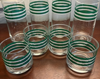 Puede incluir: Un juego de seis vasos transparentes. Tres vasos altos y tres vasos más cortos están dispuestos sobre una superficie de madera. Cada vaso presenta un diseño de tres rayas verdes horizontales.
