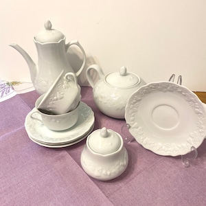 May include: A white porcelain tea set with a teapot, a creamer, a sugar bowl, cups, and saucers. The set features a raised floral pattern and is arranged on a lavender tablecloth. The teapot is approximately 10 inches tall.