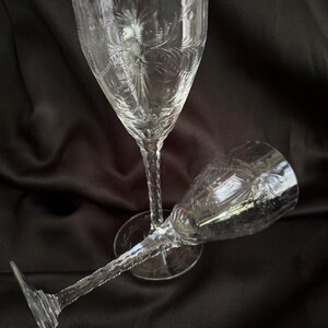 Puede incluir: Dos copas de vino de cristal transparente, grabadas con diseños florales. Una copa está en posición vertical, la otra está tumbada de lado. Los tallos tienen un aspecto texturizado y retorcido. El fondo es de color oscuro y liso.