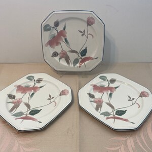 Peut inclure: Ensemble de trois assiettes octogonales en c&eacute;ramique blanche avec un motif floral. Les assiettes pr&eacute;sentent des fleurs roses, des feuilles vertes et un bord bleu fonc&eacute;. Les assiettes sont dispos&eacute;es sur une surface claire.