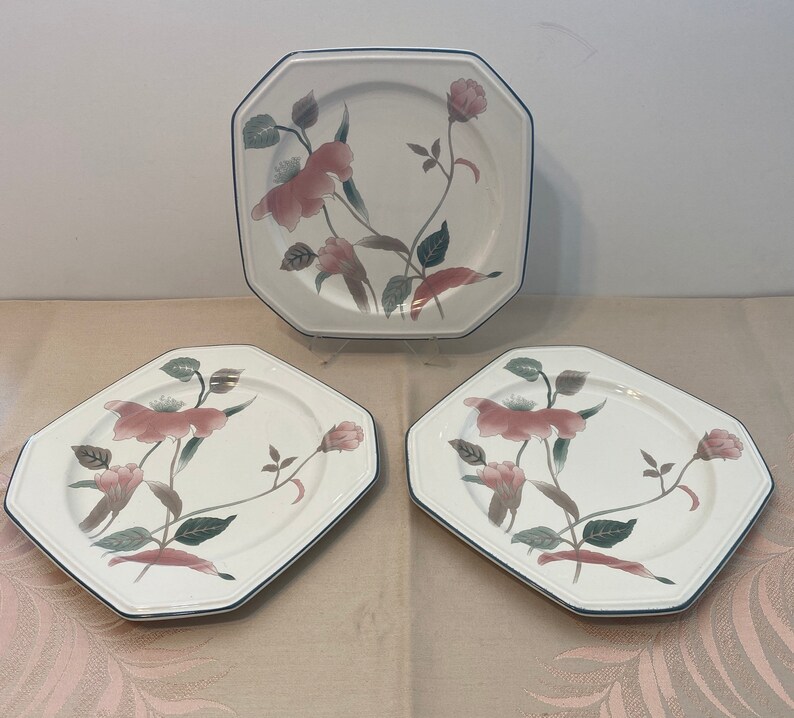 Peut inclure: Ensemble de trois assiettes octogonales en c&eacute;ramique blanche avec un motif floral. Les assiettes pr&eacute;sentent des fleurs roses, des feuilles vertes et un bord bleu fonc&eacute;. Les assiettes sont dispos&eacute;es sur une surface claire.