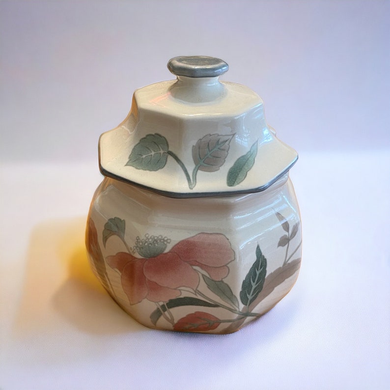 Peut inclure: Un pot en c&eacute;ramique avec un couvercle, orn&eacute; d'un motif floral dans les tons de rose, vert et marron. Le pot est de couleur cr&egrave;me, de forme hexagonale, avec un bord gris. Le couvercle est &eacute;galement cr&egrave;me avec un bouton gris.