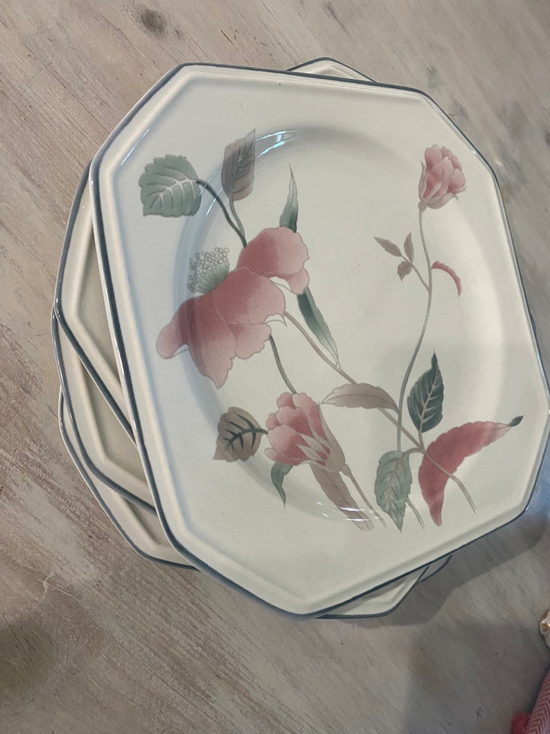Peut inclure: Une pile d'assiettes blanches octogonales avec un motif floral. Les assiettes pr&eacute;sentent des fleurs roses et marron clair avec des feuilles vertes et un bord bleu. Les assiettes sont empil&eacute;es sur une surface en bois clair.