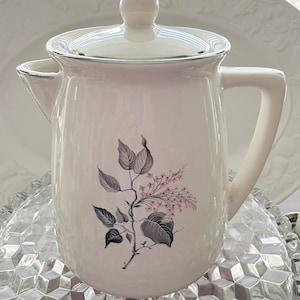 May include: Cream-coloured ceramic jug with a lid and handle. The jug features a floral design with grey leaves and pink flowers. A silver trim accents the rim and spout. The jug is displayed on a decorative glass plate.