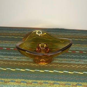 May include: An amber-colored glass candle holder with a wide, organic, free-form bowl shape and a central cylindrical socket for a single taper candle. The glass is translucent and smooth, featuring a deep golden-brown hue throughout the piece.