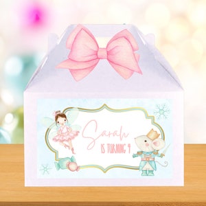 Peut inclure: Une boîte à dragées blanche avec un nœud rose sur le dessus. La boîte a une étiquette bleue et verte avec une fée ballerine, une souris en costume de soldat et le texte "Sarah a 4 ans".