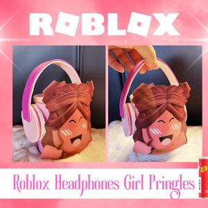 May include: A pink and white cardboard cutout of a Roblox girl wearing headphones. The girl has brown hair and is smiling. The cutout is sitting on a white surface. A can of Pringles crisps is in the lower right corner of the image.