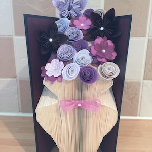 May include: A book folded into the shape of a vase with a bouquet of purple, pink, and white paper flowers. The vase is decorated with a pink ribbon and a pearl.