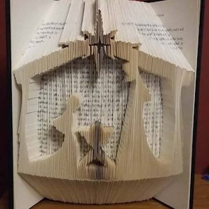 May include: A handmade book sculpture depicting a nativity scene. The pages of the book are intricately folded and cut to create the shape of a stable, with figures and a star. The book is open, revealing the detailed papercraft.