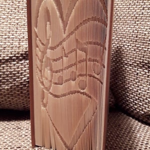 Peut inclure: Un livre décoratif dont les pages sont sculptées pour révéler un cœur et des notes de musique. Les bords des pages sont visibles et le livre a une couverture marron. Le livre repose sur une surface texturée.