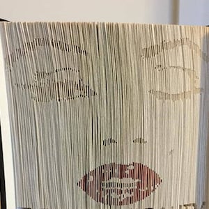 Może przedstawiać: Książka z wyciętymi stronami tworzącymi twarz. Twarz ma oczy, nos i czerwone usta. Książka ma czarny grzbiet, a strony są w jasnobeżowym kolorze.