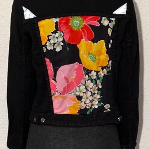 Peut inclure: Veste en jean noire avec un motif floral vibrant au dos. Le motif coloré présente des fleurs rouges, jaunes et roses avec des accents blancs. La veste a des boutons argentés et des détails réfléchissants.