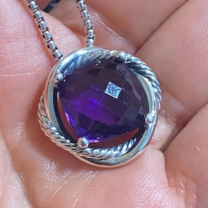 May include: A silver necklace with a large, faceted, oval-shaped, deep purple amethyst pendant. The pendant is encircled by a twisted silver rope design and a smooth silver border. The chain is a linked design.
