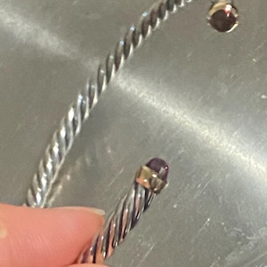 May include: A silver-toned cuff bracelet featuring a twisted cable texture throughout the band. The open ends of the bracelet are capped with small, polished gold-toned metal accents, each set with a single, round, dark purple gemstone.