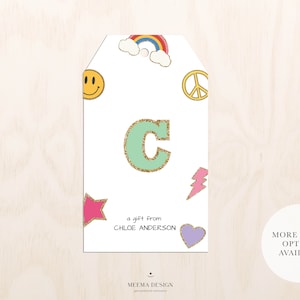 May include: A white gift tag with a green letter C with gold glitter, a rainbow, peace sign, smiley face, star, lightning bolt, and heart. The text "a gift from CHLOE ANDERSON" is printed on the tag.  "MORE COLOR OPTIONS AVAILABLE" is printed on a separate white circle.