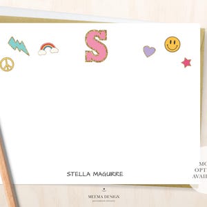 May include: A white card with a pink letter "S" and other colorful icons, including a rainbow, a lightning bolt, a peace sign, a heart, a smiley face, and a star. The card also has the text "Stella Maguire" and "Meemma Design".