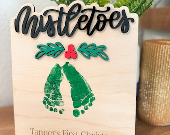 May include: A wooden sign with the words "Mistletoes" in black script. The sign has a green holly leaf and red berry design. There are green handprints on the sign with the text "Tanner's First Christmas 2023".