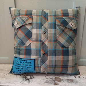 May include: Cushion made from a repurposed plaid shirt. The pillow has a blue, grey, and orange check pattern, two pockets, and button details. A blue patch with text is sewn on the front. A unique home decor item.