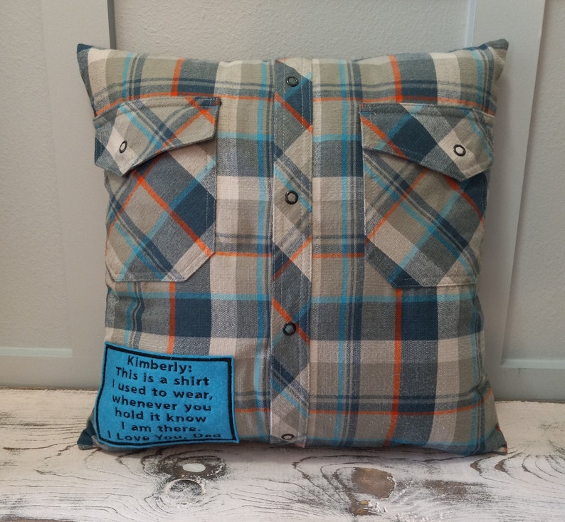 May include: Cushion made from a repurposed plaid shirt. The pillow has a blue, grey, and orange check pattern, two pockets, and button details. A blue patch with text is sewn on the front. A unique home decor item.