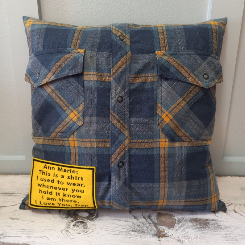 May include: Square cushion made from a blue, yellow, and grey plaid flannel shirt. The cushion has two shirt pockets and black buttons down the front. A yellow patch with text reads: "Ann Marie: This is a shirt I used to wear, whenever you hold it know I am there. I Love You, Dan."