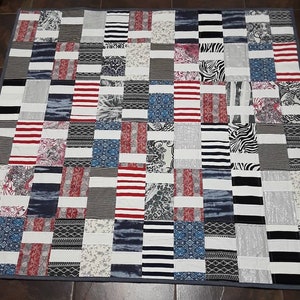 May include: A patchwork quilt with a variety of fabric patterns and colors. The quilt features rectangular blocks with stripes, floral prints, and zebra patterns in red, white, black, and blue. The quilt is bordered with a gray fabric.