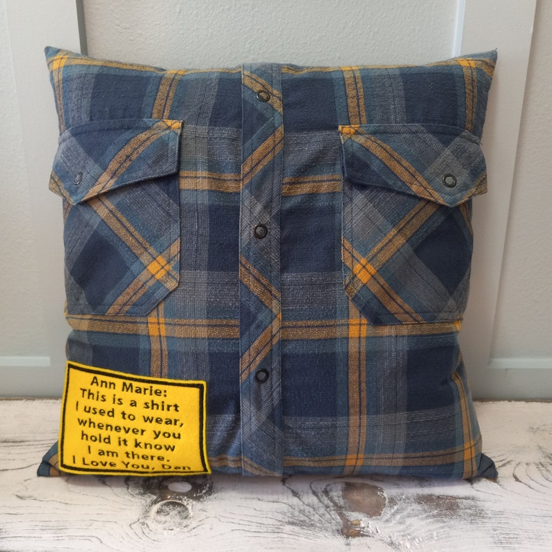 May include: Square cushion made from a blue, yellow, and grey plaid shirt. The shirt's pockets and button placket are visible. A yellow patch with text reads: "Ann Marie: This is a shirt I used to wear, whenever you hold it know I am there. I Love You, Dan."